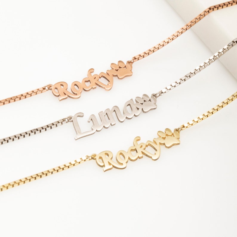 May include: Three gold, silver, and rose gold necklaces with paw print charms and personalized nameplates. The nameplates read "Rocky" and "Lumas".