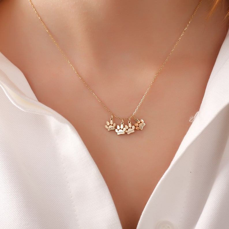 May include: Gold necklace with four small paw print charms, each engraved with a different name: Lyra, Skye, Jasmine, and Luna.