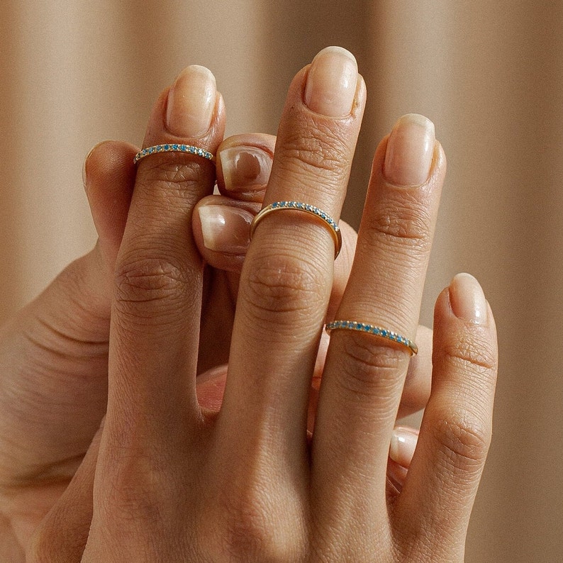 May include: A hand model wearing three thin gold rings with blue gemstones. The rings are worn on the index, middle, and ring fingers.