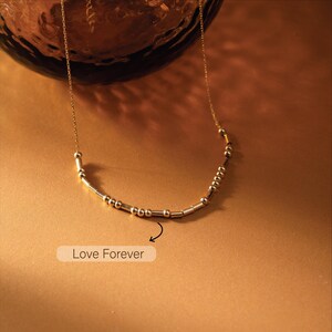 May include: A delicate gold-colored necklace with a series of small beads and cylindrical bars arranged in a curved design. The necklace features the phrase "Love Forever" in Morse code. The chain is thin and the pendant is centered.