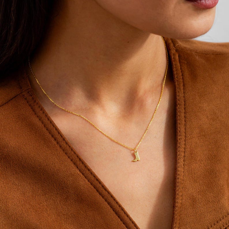 Op de afbeelding: Een delicate goudkleurige ketting met een kleine hanger in de vorm van een cowboylaars. De hanger hangt aan een fijne ketting, waardoor een eenvoudig maar stijlvol accessoire ontstaat. De ketting wordt gedragen tegen een bruine achtergrond.