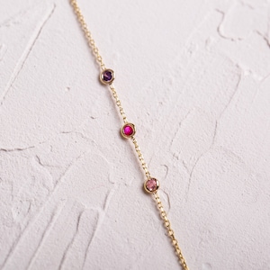 May include: A gold chain necklace with four small gemstones. The gemstones are purple, pink, red, and pink.