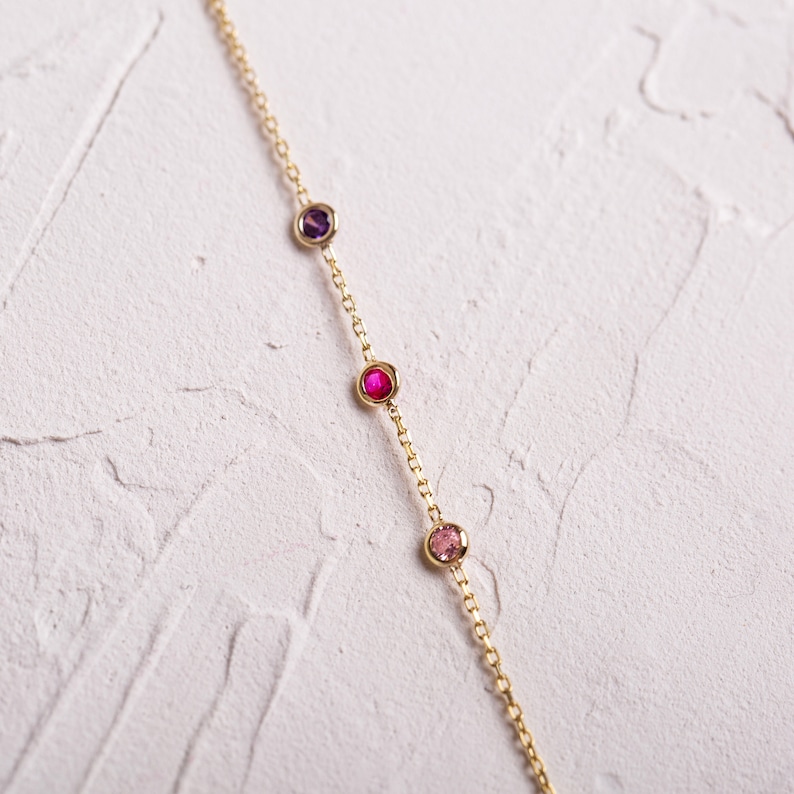 May include: A gold chain necklace with four small gemstones. The gemstones are purple, pink, red, and pink.
