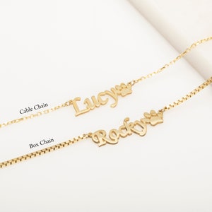 May include: Two gold necklaces with personalized names "Lucy" and "Rocky" with a paw print charm. The necklaces are on a white background. One necklace has a cable chain and the other has a box chain.