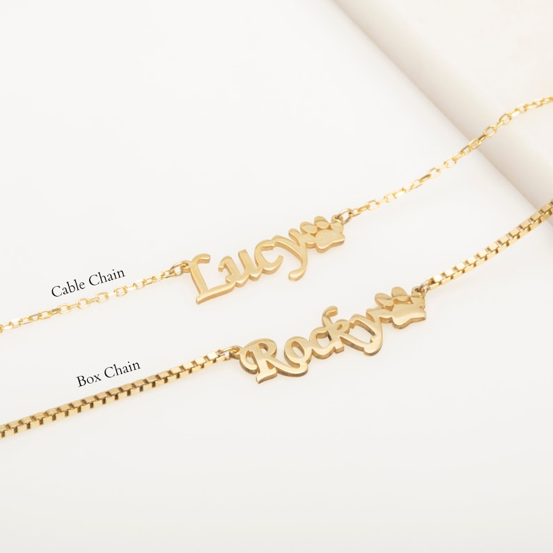 May include: Two gold necklaces with personalized names "Lucy" and "Rocky" with a paw print charm. The necklaces are on a white background. One necklace has a cable chain and the other has a box chain.