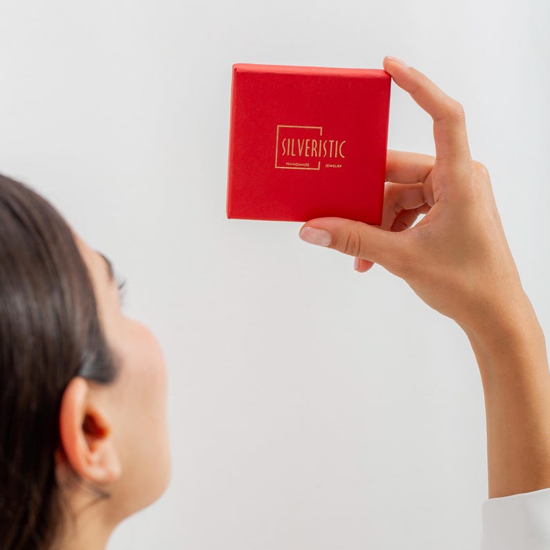 May include: A red square box with the word "SILVERISTIC" in gold lettering. The box is held up by a person's hand against a white background. The box appears to be a jewellery box.