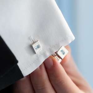 May include: A pair of silver-toned rectangular cufflinks featuring a central light blue rectangular stone inlay. The cufflinks are secured through the buttonholes of a crisp white dress shirt cuff, showcasing a polished metallic finish.