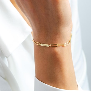 May include: A gold chain bracelet with a rectangular tag that says "SARAH" engraved on it.