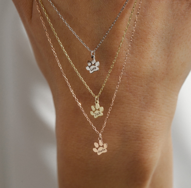 Op de afbeelding: Drie gouden halskettingen met een pootprint, met de namen "Luna", "Jack" en "Charlie" erop gegraveerd. De kettingen liggen op elkaar op de pols van een persoon.