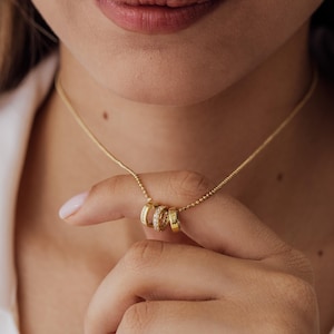 Könnte beinhalten: Eine goldfarbene Halskette mit einer feinen Kugelkette und drei kleinen runden Anhängern. Zwei der Anhänger sind glatte Goldringe, die mit den Namen Brianna und Taylor graviert sind, während der mittlere Anhänger mit klaren Steinen besetzt ist.