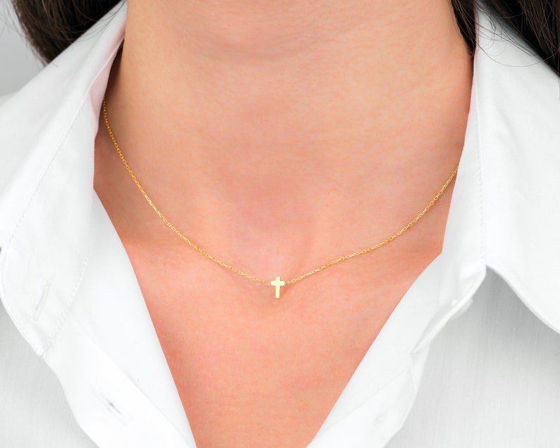 Pu&ograve; includere: Una delicata collana d'oro con un piccolo pendente a forma di croce. La croce &egrave; centrata su una sottile catena d'oro. La collana &egrave; indossata su una camicia bianca.