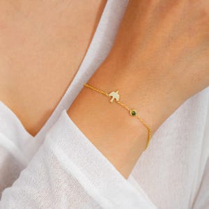 May include: A gold chain bracelet with a small bird charm and a green gemstone. The bracelet is on a person's wrist.