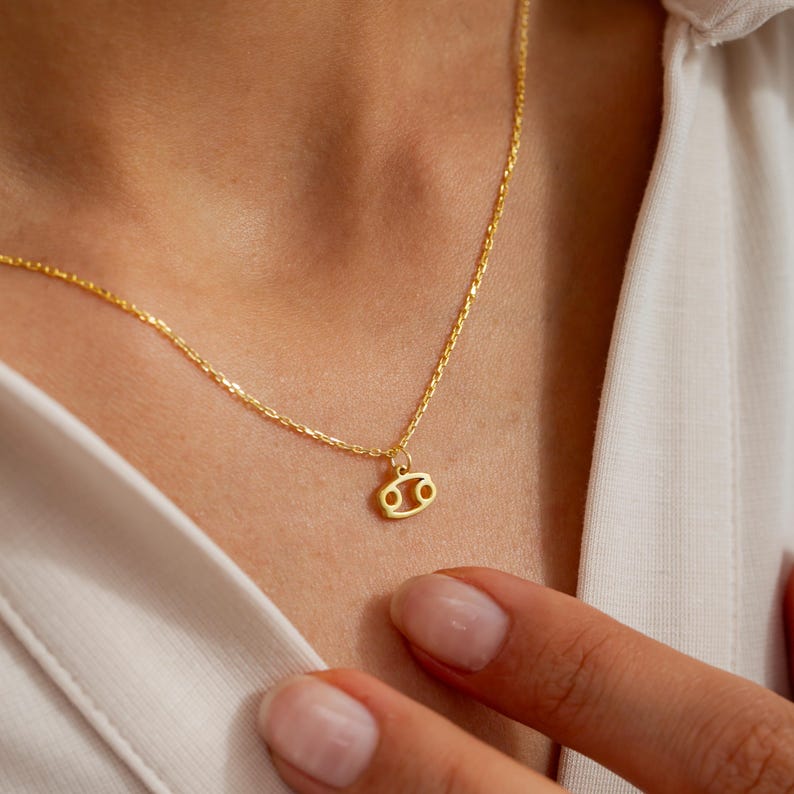 May include: A gold chain necklace with a small gold charm in the shape of the Cancer zodiac sign.