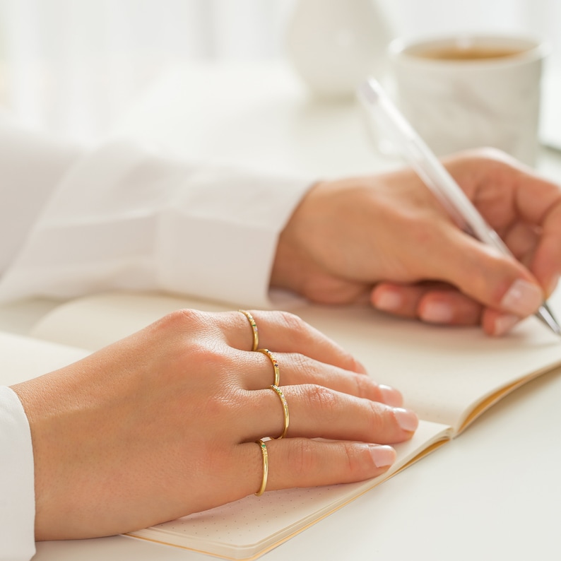 May include: Four thin gold rings worn on the fingers, each featuring a small, multi-colored gemstone accent. The rings have a smooth, polished finish and are worn stacked across the fingers, showcasing a delicate and simple design.