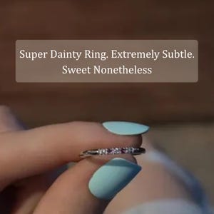 May include: A thin silver-toned ring featuring a row of small, sparkling clear stones set along the band. The text Super Dainty Ring. Extremely Subtle. Sweet Nonetheless is displayed in white font on a brown rectangular background above the ring.