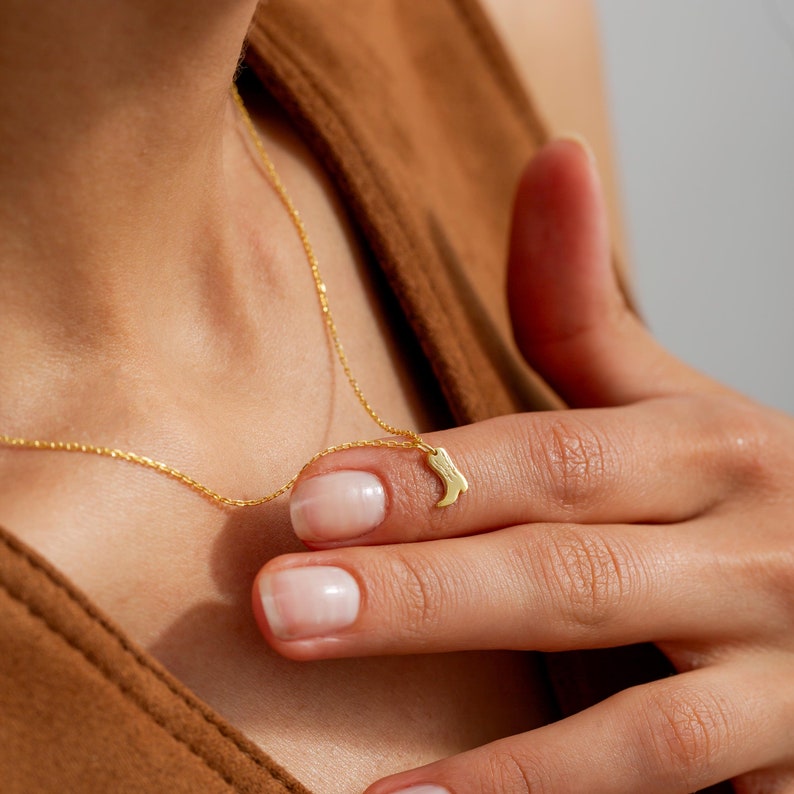 Op de afbeelding: Een delicate goudkleurige ketting met een kleine hanger in de vorm van een cowboylaars. De hanger hangt aan een fijne ketting. De ketting wordt getoond op een persoon die een bruine top draagt.