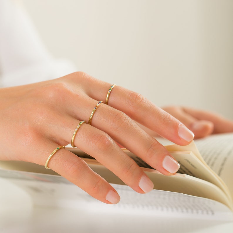 May include: Four thin gold rings worn on the fingers, each featuring a small row of tiny, multicolored gemstones. The bands are smooth and polished, with the stones set in a linear arrangement along the top section of each ring.