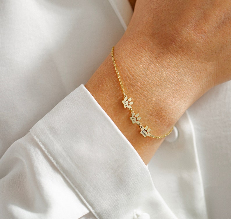May include: Gold chain bracelet with four paw print charms. Each charm has a different name engraved on it.