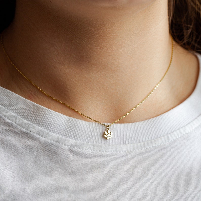 Op de afbeelding: Een gouden ketting met een klein hangertje in de vorm van een poot. De ketting wordt gedragen door iemand met een wit shirt.
