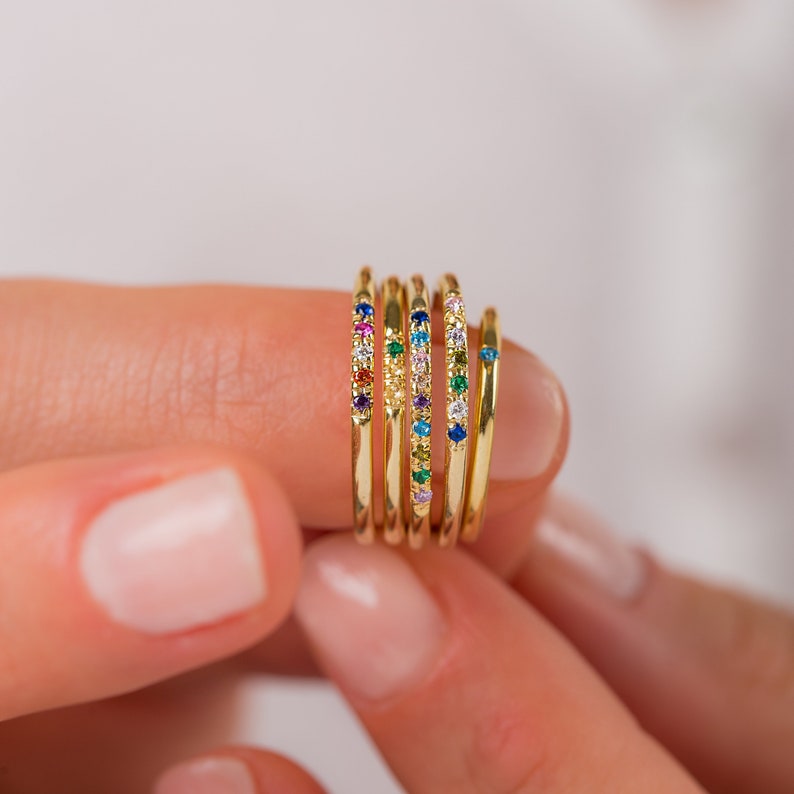 May include: A set of five thin gold rings, each featuring small, colorful embedded gemstones. The rings display various arrangements of blue, green, red, yellow, and purple stones, with one ring featuring a single blue stone.