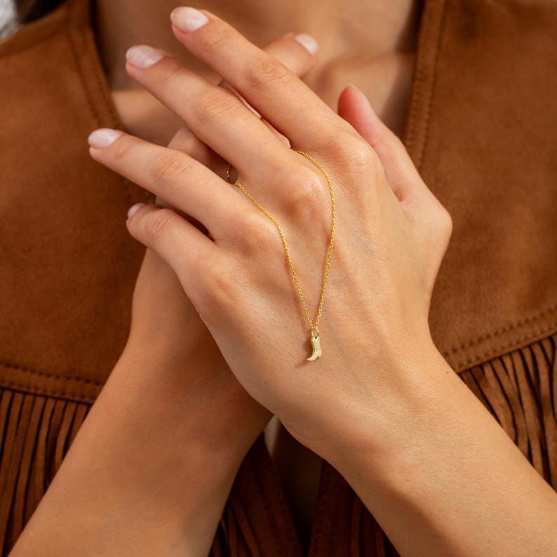 Op de afbeelding: Een delicate goudkleurige ketting met een kleine laarshanger. De ketting is dun en de hanger is een gedetailleerde miniatuur van een cowboylaars. De ketting wordt gepresenteerd tegen een bruine achtergrond.