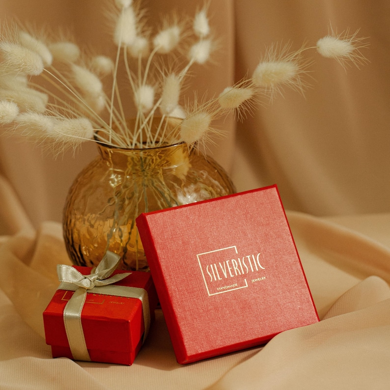 May include: A red box with the text "SILVERISTIC" in gold lettering. The box is sitting on a beige surface next to a vase filled with dried white flowers.