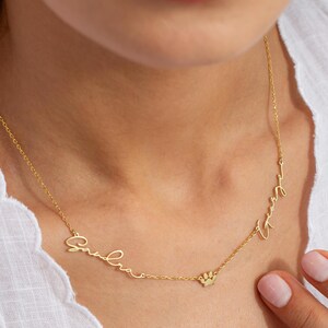 May include: Gold necklace with two names, "Gabriela" and "Wendy", written in cursive script. A small paw print charm hangs between the names.