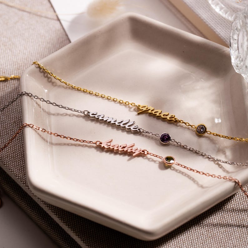 May include: Three delicate gold, silver, and rose gold necklaces with small gemstones. Each necklace features a name, "grace", "camille", and "katelyn", written in cursive.