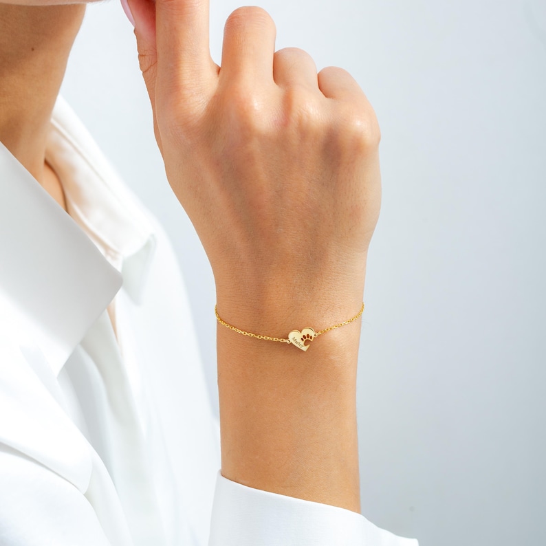 May include: A delicate gold bracelet with a heart-shaped charm, featuring a paw print design. The charm is connected to a thin chain. The bracelet is worn on a wrist. The background is a soft, neutral colour.