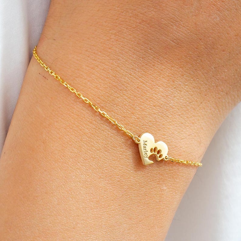 May include: A delicate gold bracelet with a heart-shaped charm. The charm has a paw print cutout and the name "Marley" engraved. The bracelet is attached to a fine chain.
