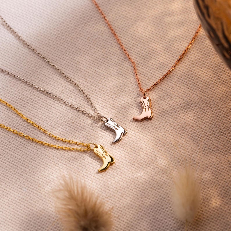 Op de afbeelding: Drie kettingen met cowboylaars-hangers in goud, zilver en ros&eacute;goud. De hangers zijn gedetailleerd met ge&euml;tste lijnen en de kettingen zijn fijn. De kettingen worden gepresenteerd op een neutrale, getextureerde ondergrond.