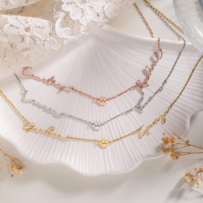 May include: Three gold, silver, and rose gold necklaces with paw print charms. Each necklace has a personalized name in cursive script. The names are "Courtney", "Lauren", "Gabriel", "Maxwell", "Thomas", and "Amy".