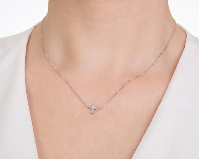 Pu&ograve; includere: Una delicata collana d'argento con un piccolo pendente a forma di croce. La croce &egrave; posizionata leggermente decentrata su una catena sottile. La collana &egrave; esposta su sfondo bianco.