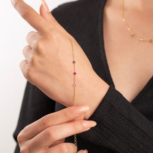 May include: A gold chain bracelet with three small gemstones: a pink, a red, and a purple.
