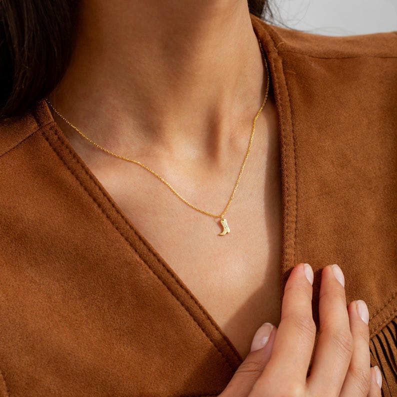 Op de afbeelding: Een delicate goudkleurige ketting met een kleine hanger in de vorm van een cowboylaars. De ketting wordt gepresenteerd tegen een bruine achtergrond, met de ketting die op de nek en borst van een persoon rust.
