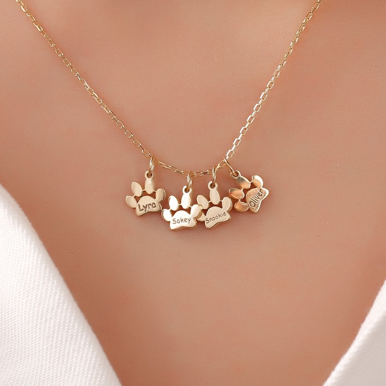 May include: Gold necklace with four paw print charms, each engraved with a different name: Lyla, Sokey, Snookie, and Oliver.