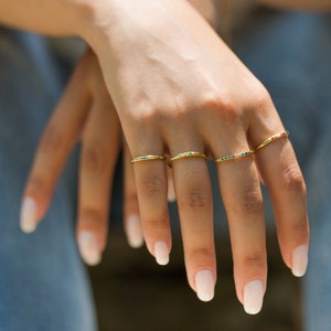 May include: Four thin gold-toned rings worn on the fingers. Each ring features a different arrangement of small, multicolored gemstones embedded along the band, including blue, green, yellow, and pink hues set into the polished metal surface.