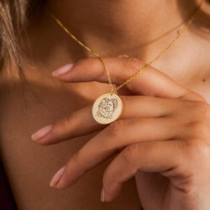 Könnte beinhalten: Eine goldfarbene Halskette mit Anhänger, die einen kreisförmigen Anhänger mit einem eingravierten Porträt aufweist. Der Anhänger hängt an einer feinen Kette. Das Porträt zeigt zwei Personen. Die Halskette wird von einer Person gehalten.