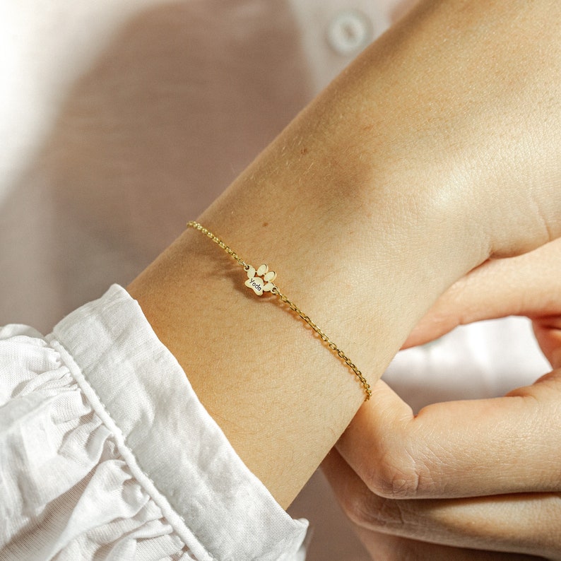 May include: A gold chain bracelet with a small paw print charm engraved with the word "Yoda".