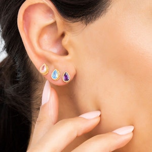May include: Three pairs of gold teardrop earrings with different colored gemstones. The earrings are shown on a person's earlobe. The gemstones are pink, blue, and purple.