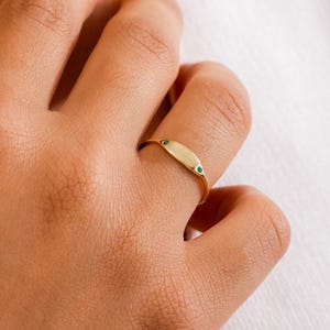 Puede incluir: Un anillo dorado con una cara ovalada adornada con dos pequeñas piedras verdes. El anillo se lleva en un dedo. Esta joya presenta un diseño sencillo y elegante.