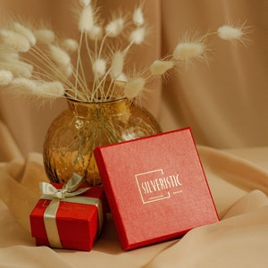May include: Two red gift boxes with gold ribbon, one with the text "SILVERISTIC HANDMADE JEWELRY" on the top, sitting on a beige surface with a vase of dried white pampas grass in the background.