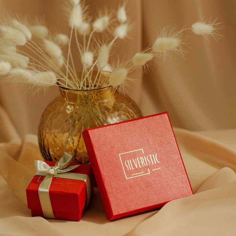 May include: A red box with the text "SILVERISTIC HANDMADE JEWELRY" on top, next to a smaller red box with a silver ribbon. The boxes are sitting on a beige surface with a vase of white fluffy flowers in the background.