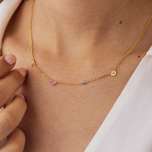 Puede incluir: Un collar de cadena dorada con cuatro pequeños dijes: una letra E, un corazón rosa, una pequeña piedra preciosa azul y un disco circular grabado con el símbolo del zodiaco Aries. Los finos eslabones dorados conectan cada dije.