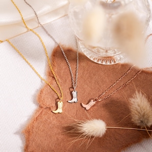 Op de afbeelding: Drie kettingen met cowboylaars-hangers in goud, zilver en ros&eacute;goud. De hangers zijn klein en gedetailleerd en hangen aan fijne kettingen. De kettingen worden gepresenteerd op een bruin oppervlak met decoratieve elementen.