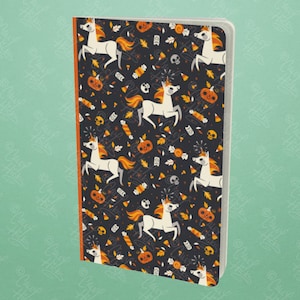 May include: A black notebook with a Halloween themed pattern featuring white unicorns, orange pumpkins, candy corn, and skulls. The notebook has a red spine.