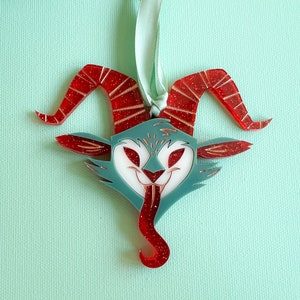 May include: Red and blue glitter ornament shaped like a horned creature with a wide, toothy grin and a long, curled tongue.