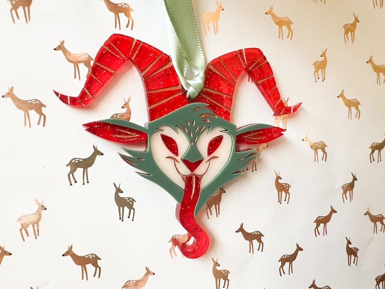 May include: A red and green Christmas ornament shaped like a whimsical creature with horns and a long tongue. The ornament is decorated with glitter and is surrounded by a white background with gold deer silhouettes.