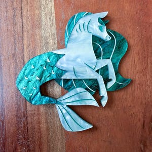 May include: A decorative brooch shaped like a hippocampus, a mythological creature with the upper body of a horse and the lower body of a fish. The piece features a pearlescent white horse section and a deep emerald green, marbled fish tail.