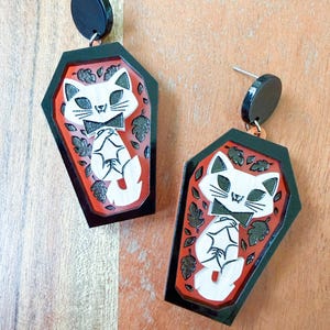 May include: A pair of dangle earrings featuring a black coffin-shaped frame with an orange background. Inside each coffin is a white cat with a black bowtie, surrounded by small black leaf motifs. The earrings have a black circular stud top.